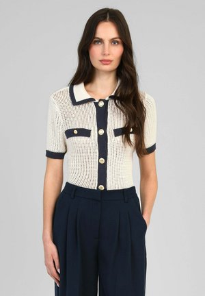 Femme portant un haut en maille blanche à manches courtes avec des bordures et des boutons bleu marine, associé à un pantalon plissé bleu marine taille haute, debout devant un fond gris.