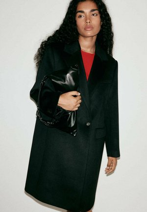 Mujer con cabello largo y rizado, vestida con un abrigo negro y una blusa roja, sosteniendo un gran bolso de cuero negro sobre un fondo liso.