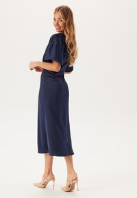 Námořnické saténové midi šaty s krátkými rukávy, přiléhavým pasem a rovnou sukní. V kombinaci s nahými sandály na podpatku. Hladká textura a jednoduchý design.