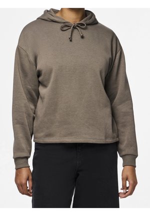 Person trägt einen taupefarbenen Kapuzenpullover mit Kordeln und schwarze Hosen, stehend vor einem schlichten weißen Hintergrund.