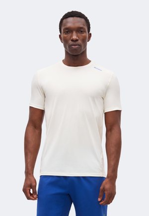 Muškarac stoji okrenut naprijed, obučen u pristajuću bijelu košulju kratkih rukava s plavim kratkim hlačama na jednobojnoj svijetloj pozadini.