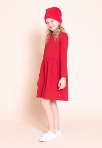 Młoda dziewczyna z kręconymi blond włosami, ubrana w czerwoną sukienkę z długim rękawem, pasującą czerwoną czapkę i białe trampki, stojąca na tle jasnego tła.