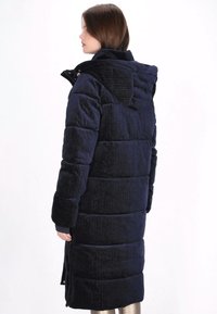 Długa, granatowa kurtka puchowa z pikowanego materiału o fakturowanej powierzchni. Posiada kaptur oraz zapięcia na zatrzaski z przodu.