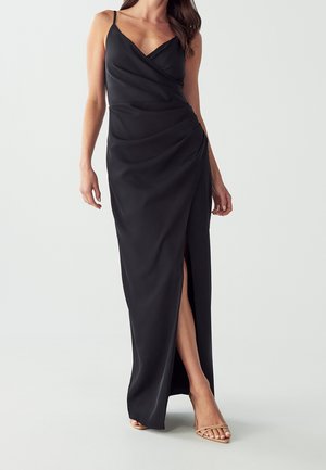 Frau trägt ein ärmelloses schwarzes Wickelkleid mit hohem Schlitz und nude-farbenen Riemchenabsätzen vor einem schlichten Hintergrund.