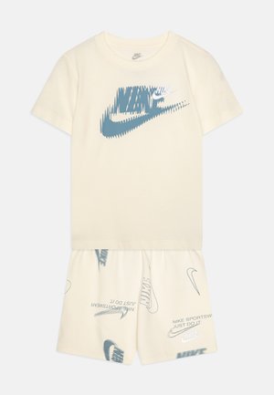 Conjunto de duas peças em bege claro, composto por uma t-shirt de mangas curtas com um logótipo gráfico azul e calções combinando com padrões do logótipo repetidos.