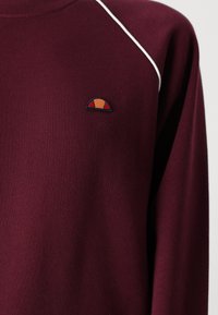 Bordeaux sweatshirt med hvid piping, der har et lille broderet logo med orange og gule accenter, lavet af et blødt, tekstureret stof.
