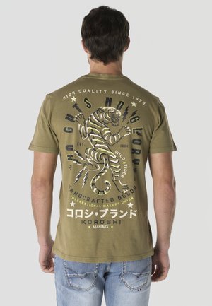 Hombre con camiseta verde oliva con gran gráfico de tigre y texto en la espalda, combinado con vaqueros azul claro, de espaldas.