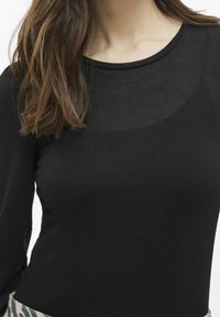 Svart långärmad topp med rund halsringning, tillverkad av slät tyg. Har en genomskinlig panel vid bröstområdet och en kontrasterande mönstrad nederkant.