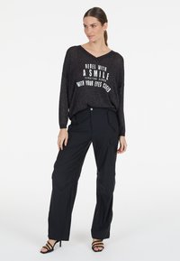 Schwarzer, übergroßer Pullover mit dem Text "REBEL WITH A SMILE", kombiniert mit schwarzen Cargo-Hosen mit Taschen und lockerer Passform. Modell trägt hohe Sandalen.