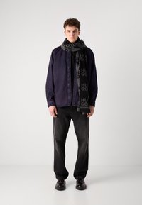 Cămașă bleumarin cu butoni, tricou negru, eșarfă gri cu logo, blugi negri drepti și pantofi negri. Un outfit simplu și modern.
