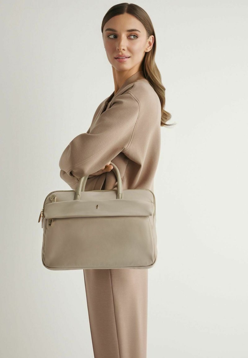 Kobieta w beżowym stroju, trzymająca taupe skórzaną teczkę, patrząca na bok na jednolitym jasnym tle.