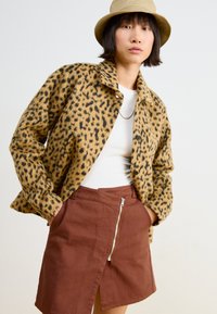 Hnědá bunda s leopardím potiskem a texturou, s knoflíky, v kombinaci s hnědými sukněmi se zipem v přední části a rozparkem, nošená přes bílý top.