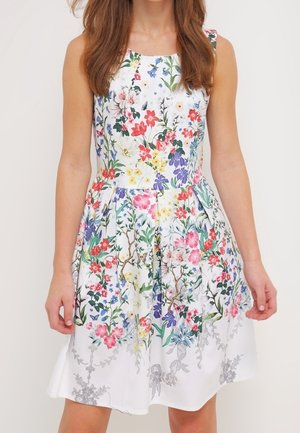 Hihaton valkoinen mekko, jossa värikäs kukka- ja kasvikuvio, vartalonmyötäinen yläosa ja laskostettu hame, pukeutuneena pitkät ruskeat hiukset omaava henkilö.