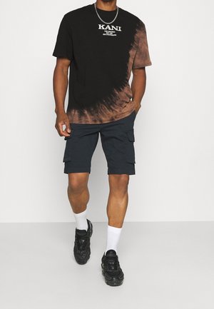 Mężczyzna ubrany w czarno-brązowy T-shirt tie-dye z białym napisem, granatowe szorty cargo, białe skarpetki, czarne trampki i srebrny naszyjnik z łańcuszkiem.