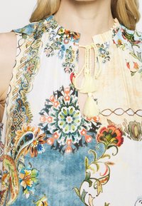 Top sin mangas floral que presenta una combinación de colores en azul y crema, con patrones intrincados, detalle de lazo con borlas en el escote y tela fruncida.