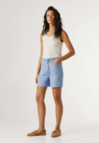 Woman stands with hand in pocket wearing white sleeveless top, light blue high-waist shorts, and brown moccasin shoes on plain background.