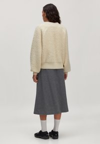 Romige gebreide trui met geribbelde zoom, gecombineerd met een grijze midi rok. Model draagt witte sokken en zwarte schoenen. Van achter gefotografeerd.
