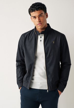 Junger Mann mit lockigem Haar, der eine schwarze Jacke mit Reißverschluss über einem hellgrauen Henley-Shirt trägt, steht mit den Händen in den Taschen einer marineblauen Hose.