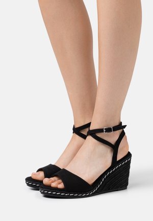 Nogi w czarnych sandałach na koturnie z paskami wokół kostki i odkrytymi palcami, z białym przeszyciem na krawędzi podeszwy, na jednolitym tle.