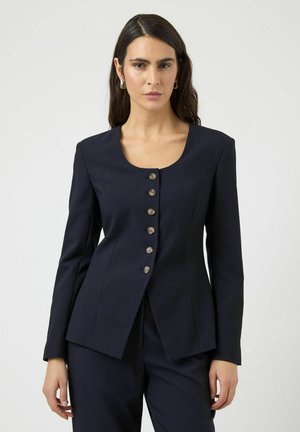 Pikkade tumedate juustega naine, kes kannab kuue nupuga tihedat mereväesinist jopet ja sobivaid mereväesiniseid pükse, seistes ühevärvilise hele tausta ees.