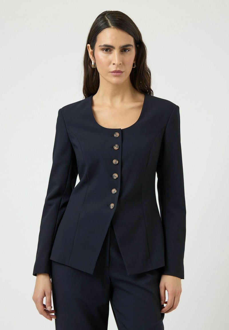 Femme aux longs cheveux foncés portant une veste cintrée bleu marine à six boutons et un pantalon assorti, debout devant un fond clair uni.