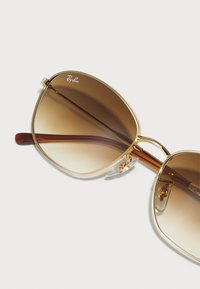 Goldene Metallsonnenbrille mit braunen farbverlaufenden Gläsern, die eine gewagte, kantige Form und klare Nasenpolster aufweist. Die Bügel sind braun mit glänzender Oberfläche.
