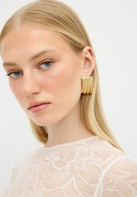 Orecchini in metallo color oro con un design ondulato, caratterizzati da scanalature sovrapposte. Il modello ha i capelli lunghi e biondi ed indossa un top in pizzo trasparente.