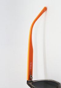 Aurinkolasit mustalla kehykset, tummilla linsseillä ja oranssilla sankalla. Sankassa on valkoiset tekstit "TIKA". CE-merkintä näkyvissä.