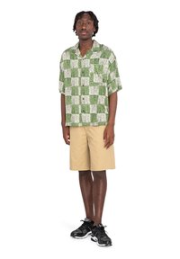 Camisa de cuadros verde y blanca de manga corta, pantalones cortos caqui y zapatos deportivos negros. La camisa tiene un corte relajado y un patrón texturizado.