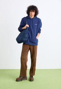 Navy blue hoodie with front pocket, paired with brown wide-leg pants and brown chunky boots. Carrying a patterned blue tote bag.