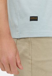Lichtblauw katoenen t-shirt met een zwart-oranje label 'READY TO DISCOVER' op de zijnnaad, gecombineerd met een beige stoffen onderkant.