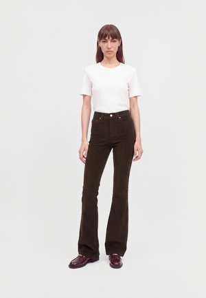 Femme aux cheveux bruns raides portant un t-shirt blanc uni à manches courtes, un pantalon évasé taille haute foncé et des chaussures bordeaux foncé, debout.