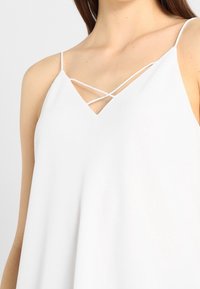 Valkoinen hihaton toppi, jossa on V-pääntie ja ristikkäiset olkaimet, valmistettu kevyistä materiaaleista, löysästi pukemiseen suunniteltu malli.