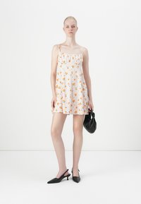 Rochie mini florală cu bretele subțiri, bază albă ornamentată cu flori portocalii, talie strânsă, tiv evazat. Modelul ține o geantă de mână neagră.