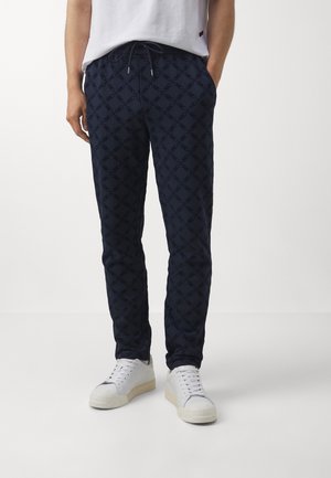 Homme portant un pantalon de jogging bleu marine à motifs, des baskets blanches et un T-shirt blanc, debout avec les mains dans les poches sur un fond blanc.