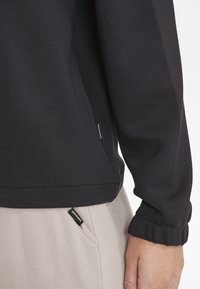 Svart sweatshirt med ribbad mudd, mjuk textur och sidobranding, ihopparad med ljusa byxor med synlig ficketikett.