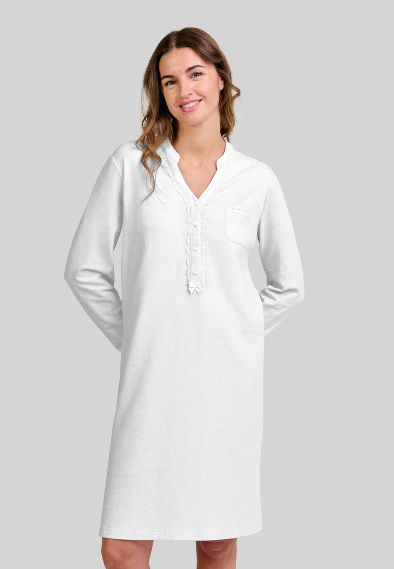 Mujer sonriendo vestida con un camisón blanco de longitud hasta la rodilla y mangas largas con botones, encaje en los bordes y un pequeño bolsillo en el pecho, sobre un fondo gris.