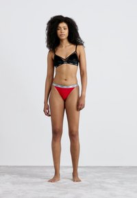 Black velvet bralette with thin straps and logo band; red low-rise thong with grey branded waistband; smooth textures, fitted design.