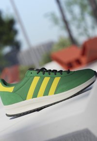Green sneaker with yellow stripes and black laces resting on a white surface with blurred outdoor background.