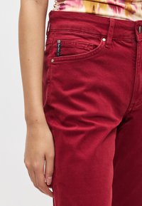 Punaiset farkut, jotka ovat kapealahkeiset ja joissa on etutaskut, metalliosat ja sileä pinta. Lähikuva vyötärön keskiosasta.