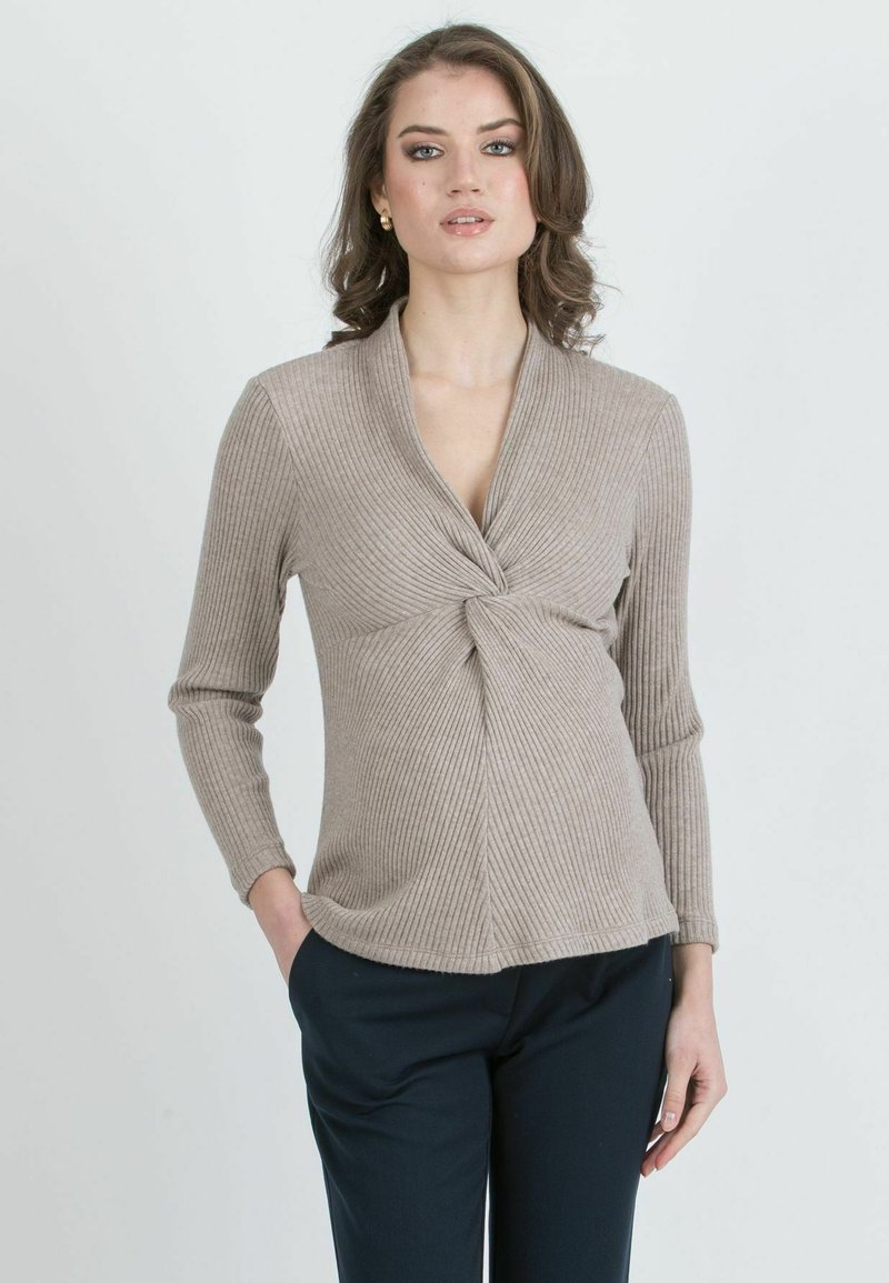 Eine Frau mit lockigem braunen Haar trägt ein beiges geripptes Langarmtop mit einem Front-Knoten und marineblauen Hosen und steht vor einem schlichten Hintergrund.