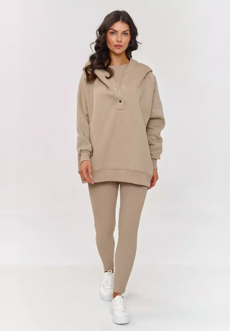 Beżowa, oversize bluza z kapturem, z przodu z klapą i guzikami, w połączeniu z beżowymi legginsami o prążkowanej fakturze i białymi sneakersami na neutralnym tle.
