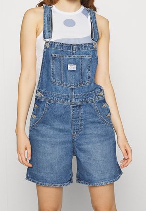 Denim tuinbroek met verstelbare bandjes, voorzakken en metalen bevestigingen. De stof heeft een middentoon blauwe wassing en een gladde textuur.