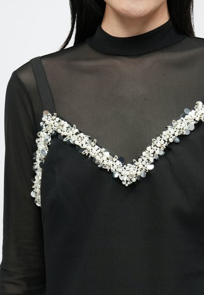 Melns apģērbs ar caurspīdīgu melnu augšdaļu, kurai ir V veida izgriezums, rotāts ar dekoratīvām pērlītēm un spīdumiem gar malām.