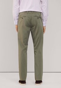 Pantaloni verde oliva con taglio dritto, due tasche posteriori con bottoni, indossati con una camicia a righe rosa chiaro e scarpe scure.