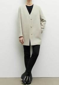 Ljust grå ullblandad rock med V-hals, tre knappar och sidofickor, tillsammans med svarta slim-fit-jeans och ankelskor.