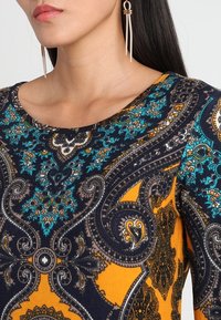 Chemise à manches longues présentant un motif cachemire en bleu, jaune et noir avec un col arrondi. Texture de tissu doux avec des détails sophistiqués.