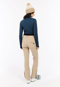 Vrouw in beige skibroek, blauwe langhuids top, beige gebreide muts met pompon, en witte skischoenen, staande met de rug naar de camera op een witte achtergrond.