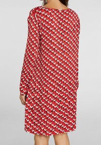 Robe rouge à manches longues, ornée d'un motif géométrique de formes blanches et rouges plus foncées. Le tissu a une texture lisse et un design plissé.