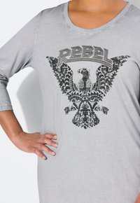 Graues Baumwollhemd mit 3/4-Ärmel, das ein schwarzes Druckdesign eines Adlers und das Wort "REBEL" darüber aufweist. Weicher Stoff, lockere Passform.